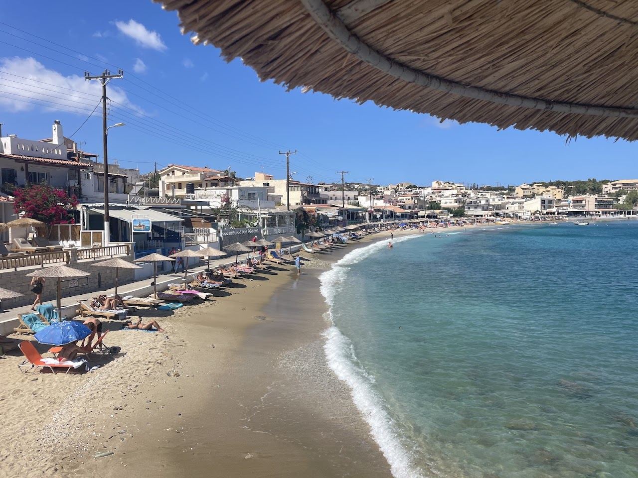 Curve of sand, straw umbrellas and clear turquoise water at the Agia Pelagia cove in Crete, with village houses reaching the shore