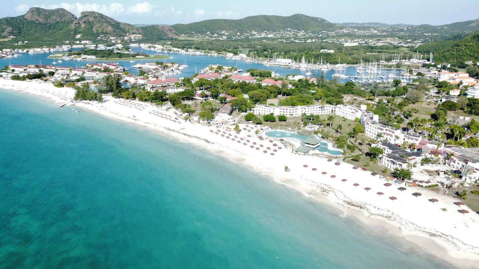 A long calm strip of turquoise water and pale sand lined with beach life on the west coast of Antigua