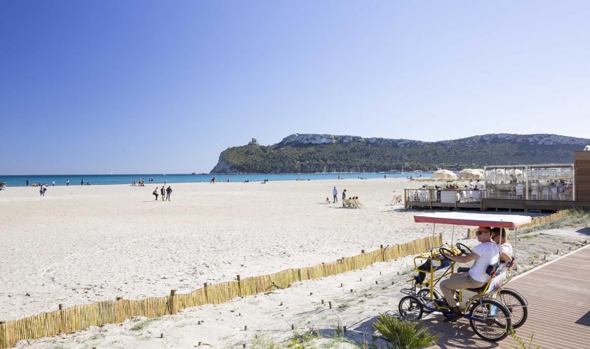 A long straight beach of pale sand and bright blue water with a green headland at Poetto in Cagliari, Sardinia