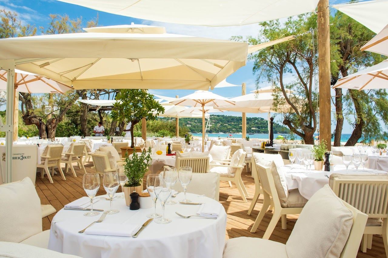 White parasols over set lunch tables on the pale timber deck at Loulou Ramatuelle with tamarisk trees and Pampelonne bay behind