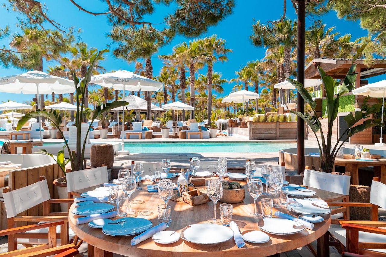 A set dining table beside the turquoise pool at Nikki Beach Saint Tropez with white day beds and parasols under the palms behind