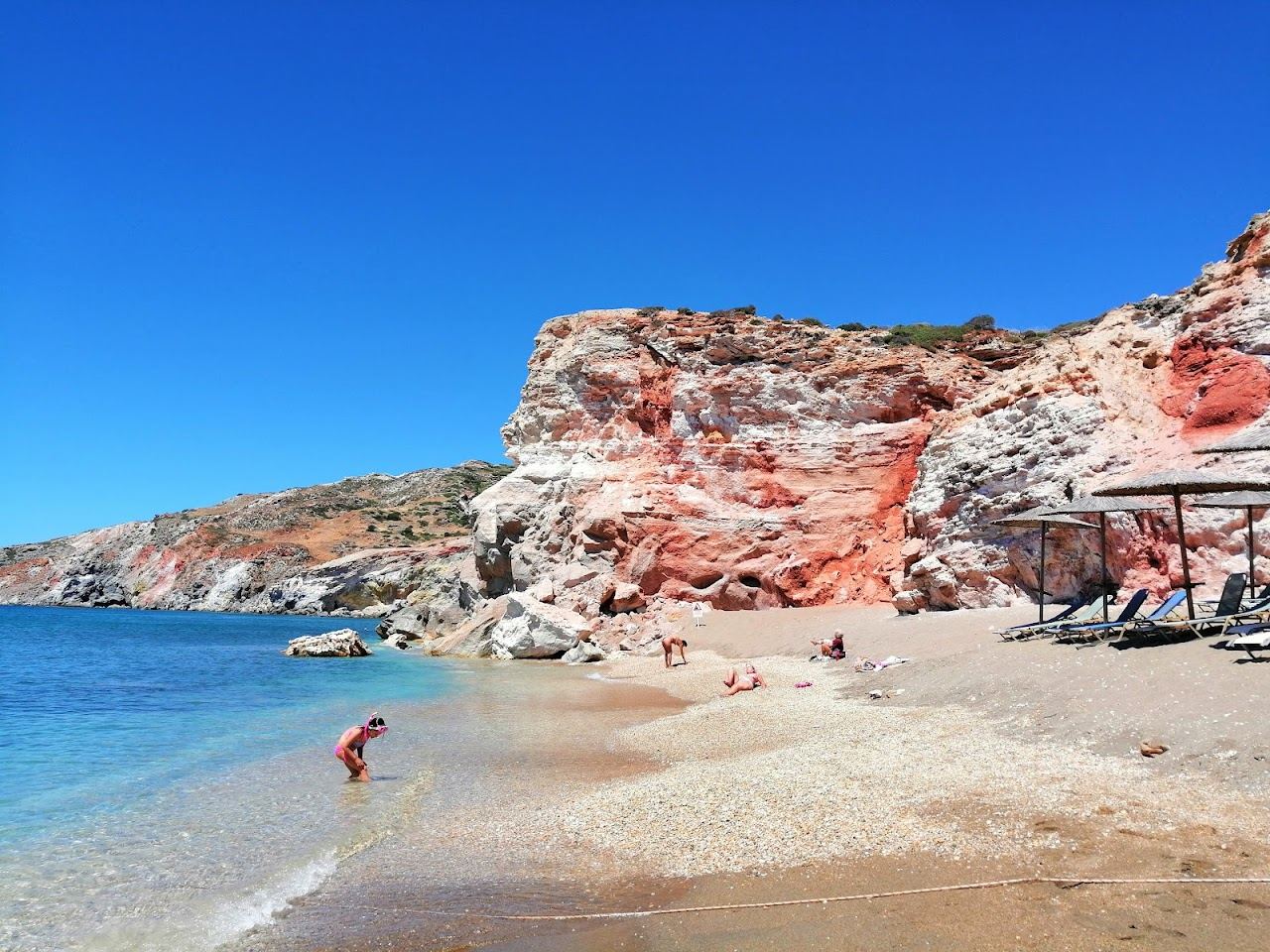 The broad beach of Paliochori below vivid streaked volcanic cliffs with sunbeds and clear water on the south coast of Milos